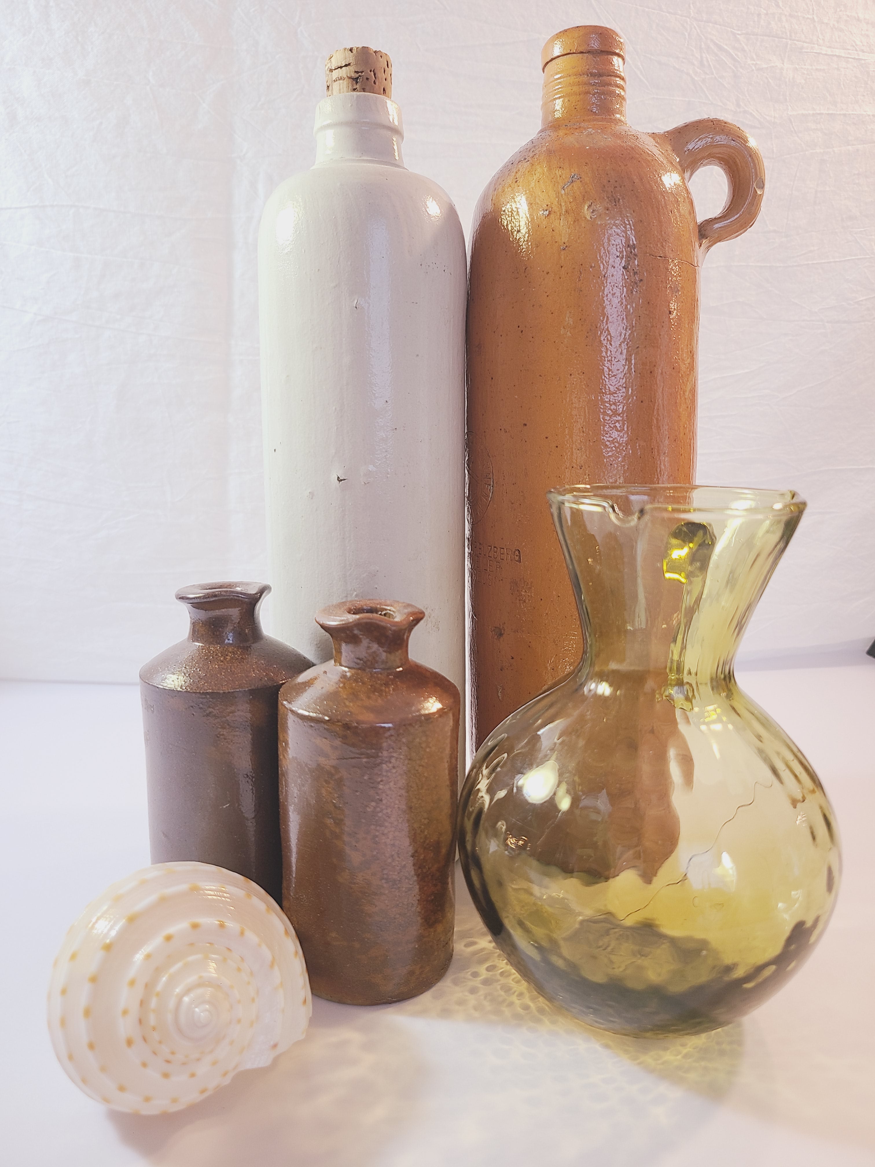 Antique 19th century terracotta colored stoneware bottle stamped with the name of Georg Kreuzberg and the Apollinaris Brunnen displayed with other stoneware bottles, glass pitcher, and seashell.