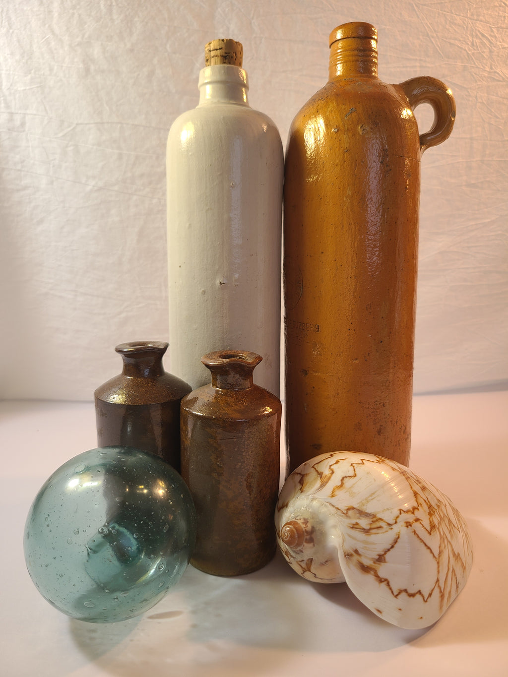 Antique 19th century terracotta colored stoneware bottle stamped with the name of Georg Kreuzberg and the Apollinaris Brunnen displayed with glass fishing float, seashell, and other stoneware bottles.