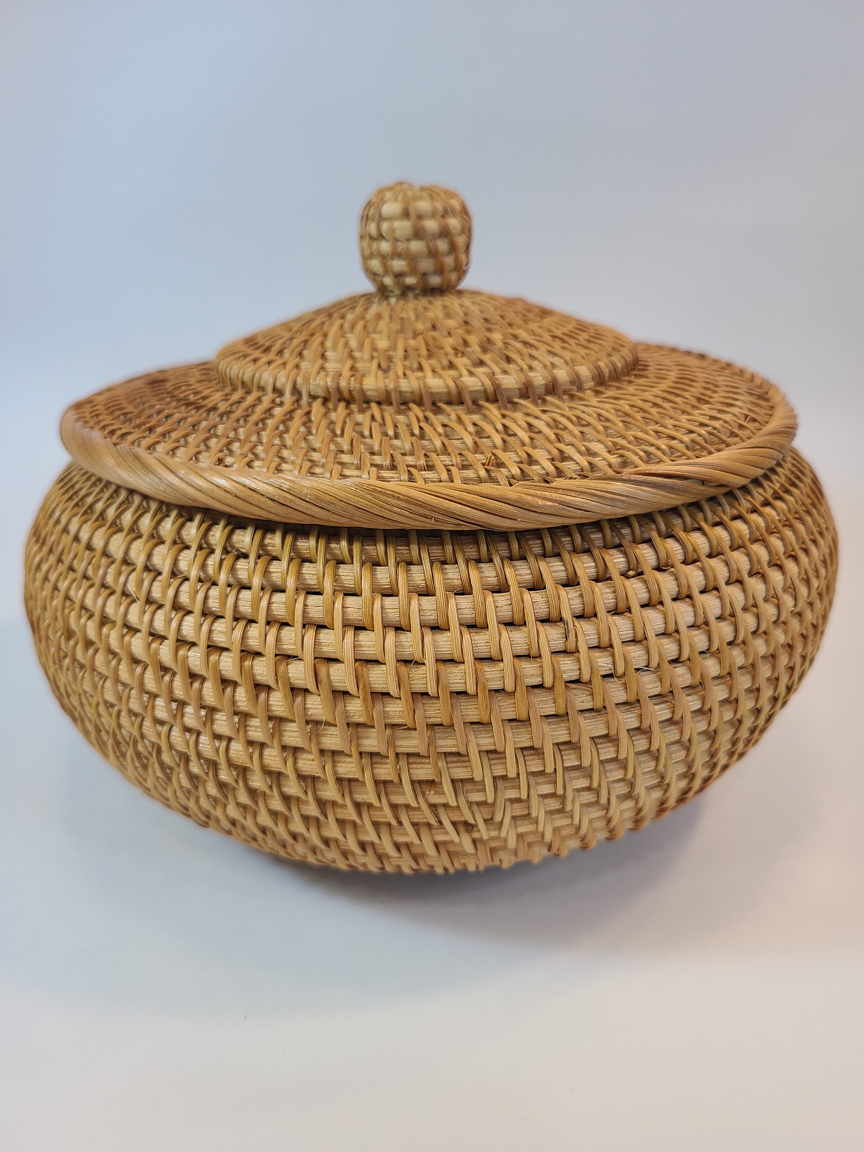 A round woven basket with a matching lid, photographed against a plain light background. The basket is made from natural tan fibers with a tightly woven pattern and a small knob on the lid.