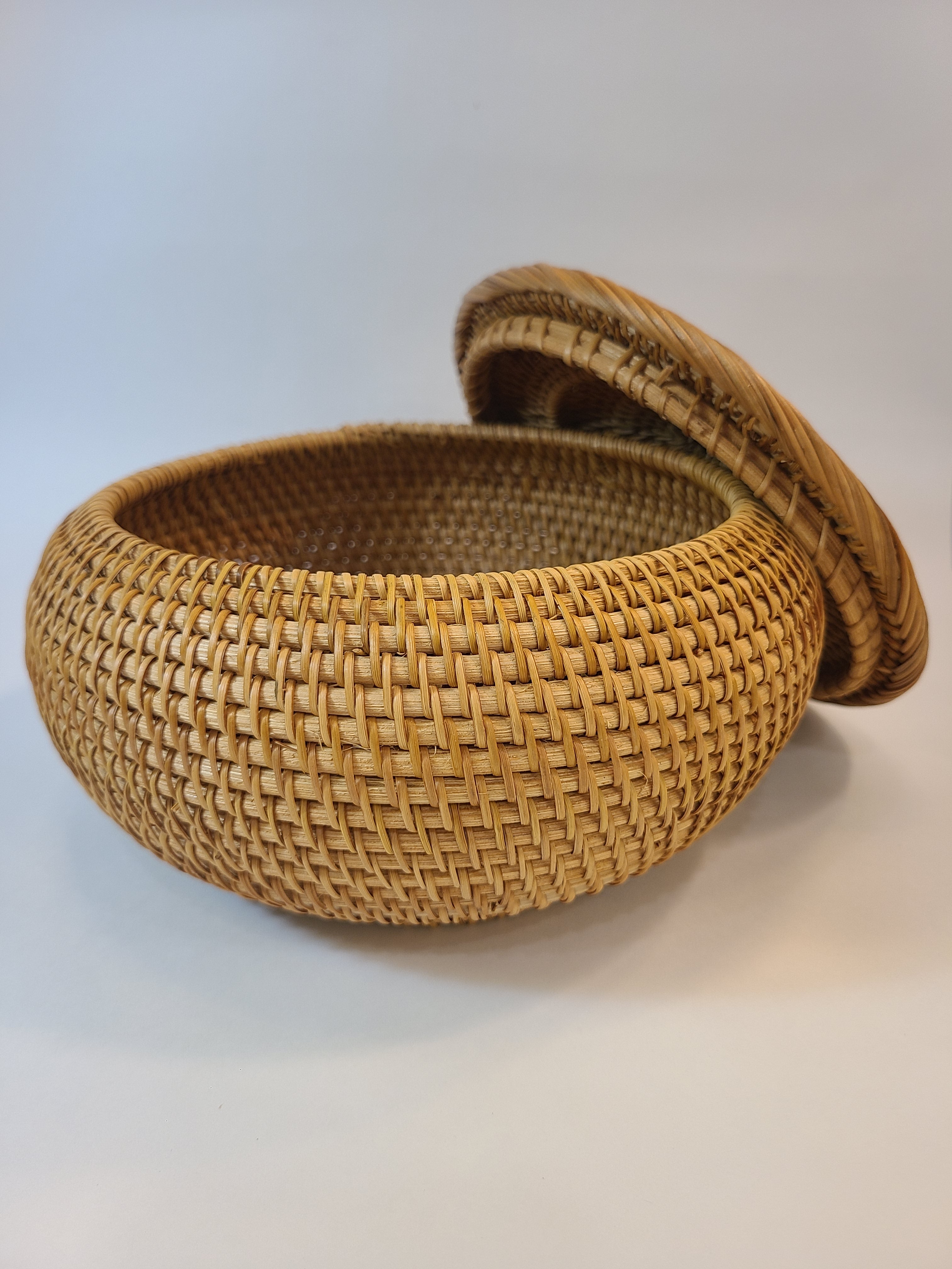 A round woven basket with a matching lid, made from natural light brown reed or straw fibers, photographed on a neutral light background.