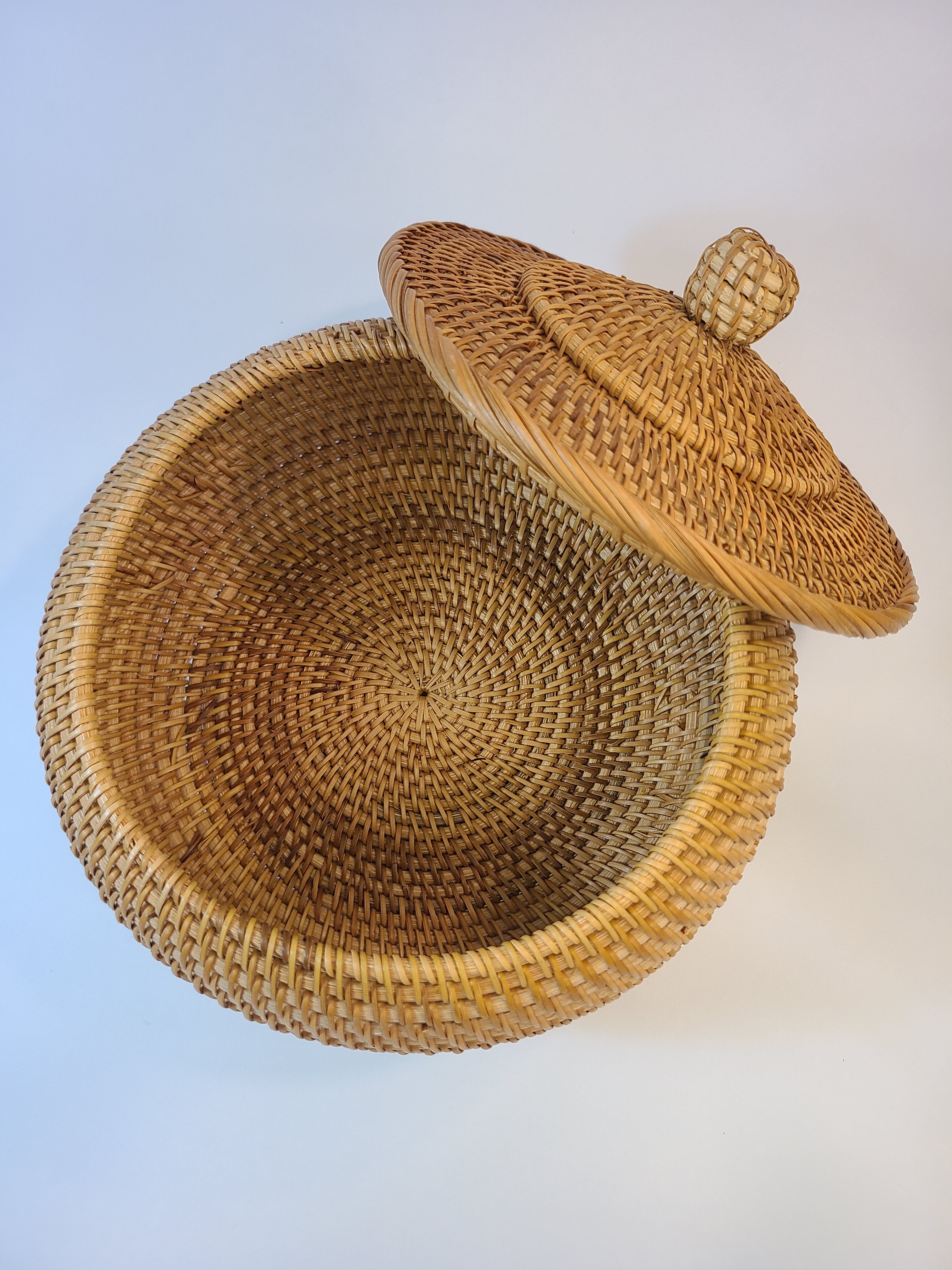 A round woven basket with a matching lid, shown open against a plain light background. The basket is made from natural tan and brown woven fibers, with a small decorative finial on top of the lid.