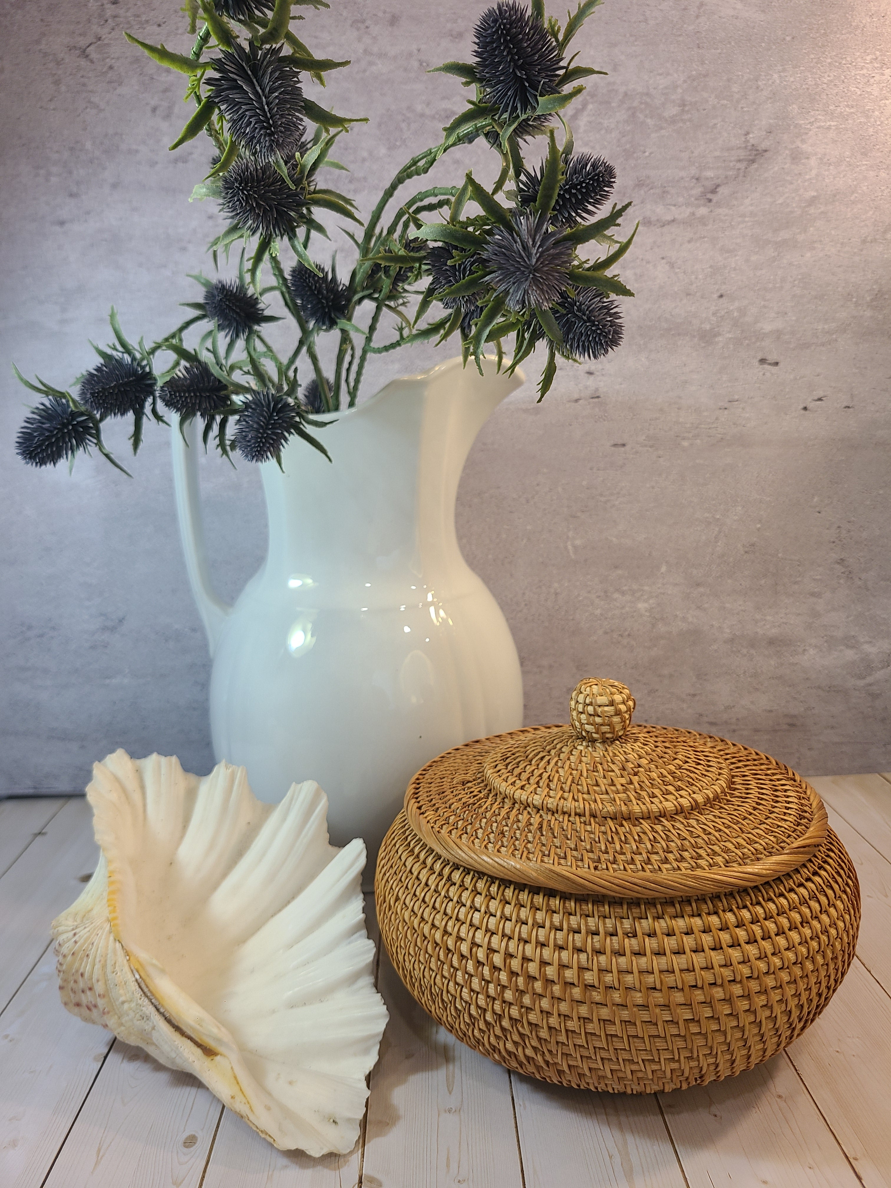A natural woven rattan storage basket with a matching lid sits on a light wood floor next to a large white seashell and a white ceramic pitcher holding ephedra plants.