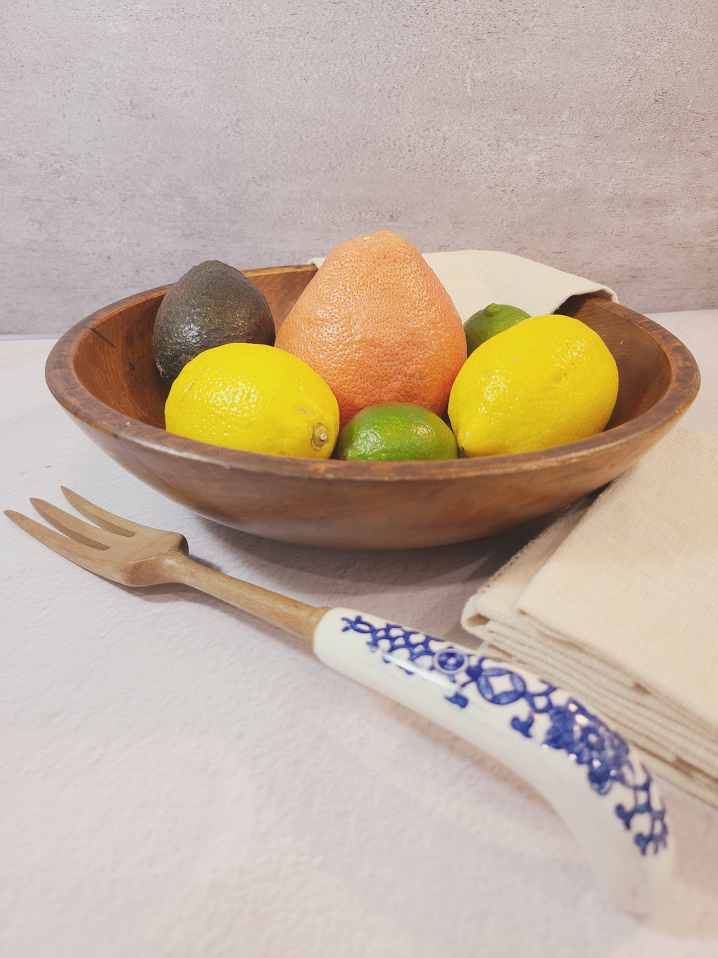 A vintage wooden bowl filled with fruit, including lemons, limes, and a grapefruit, with a decorative white and blue ceramic serving fork resting beside it on a light fabric surface.