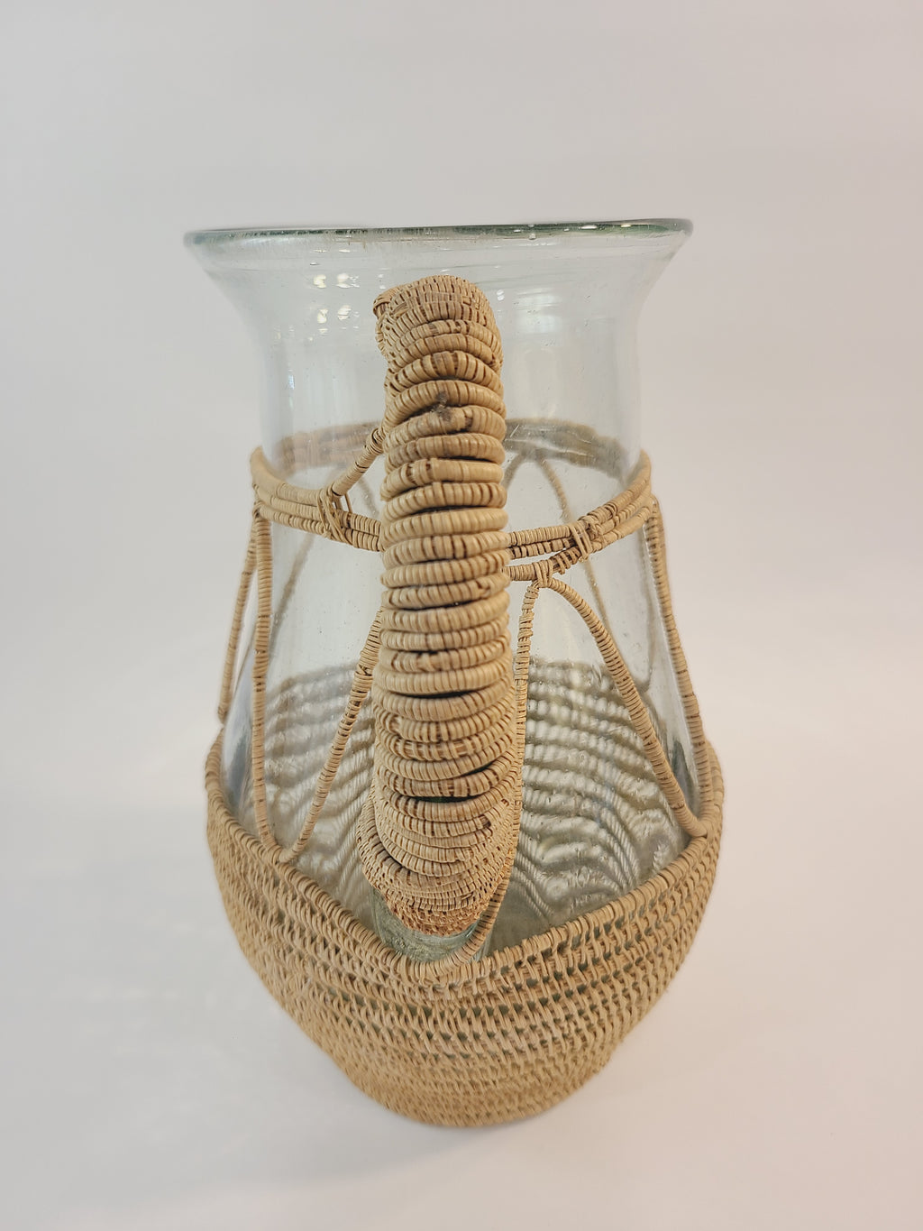 A vintage clear glass pitcher wrapped in a woven natural fiber photographed to highlight the handle.