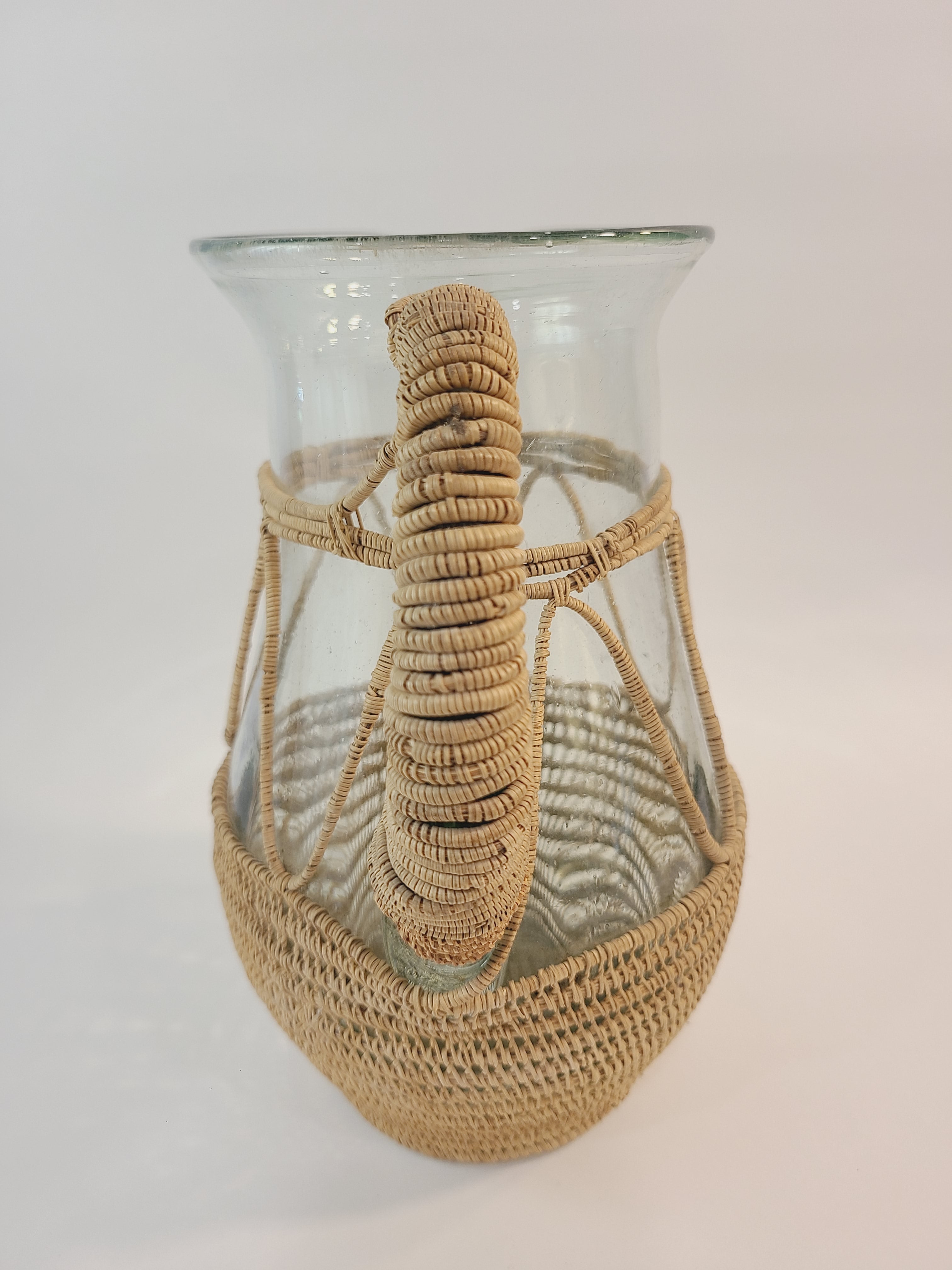 A vintage clear glass pitcher wrapped in a woven natural fiber photographed to highlight the handle.