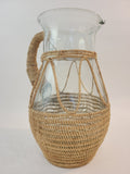 A vintage clear glass pitcher wrapped in a woven natural fiber shown on a plain light background. 