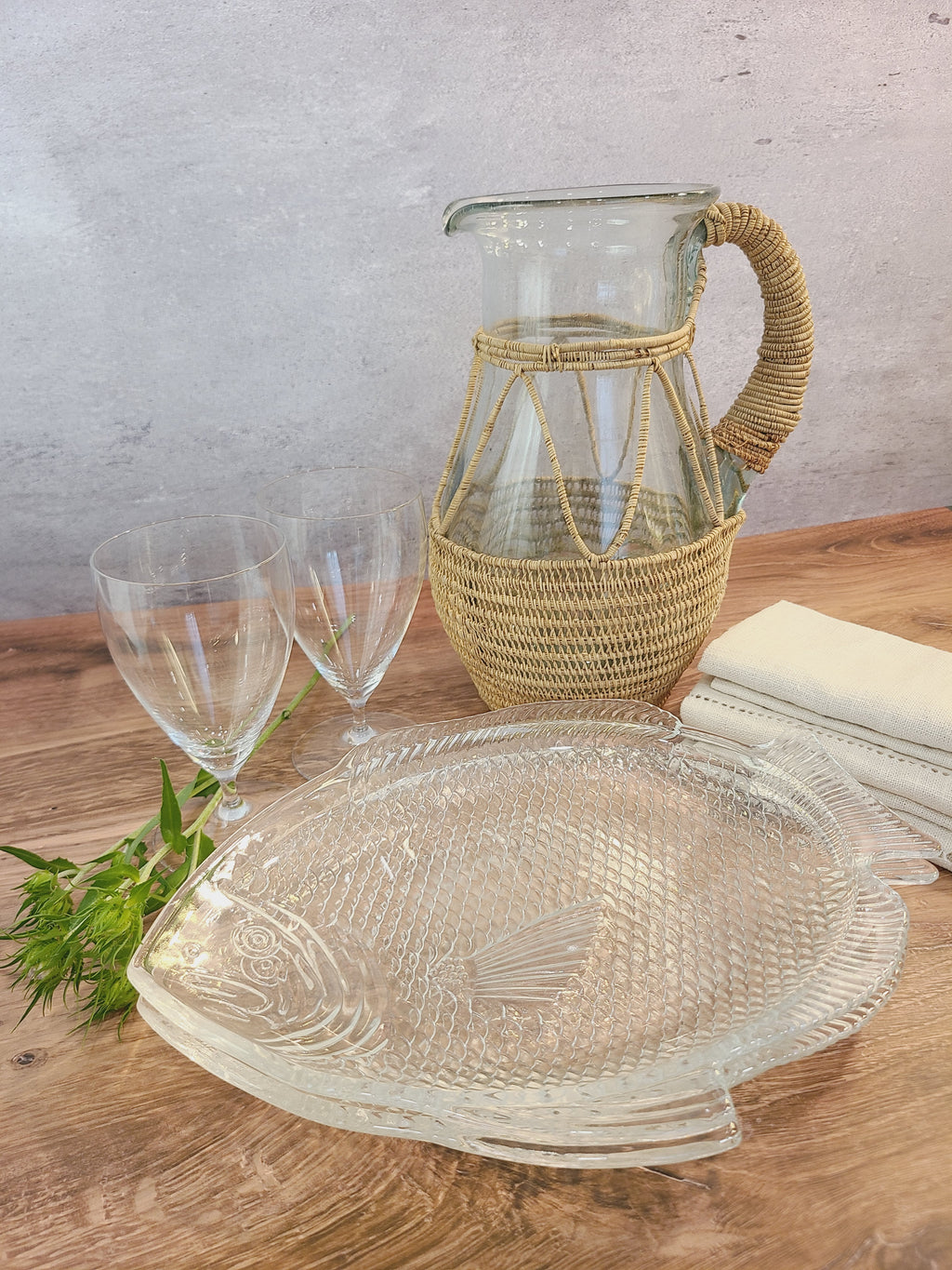 A vintage clear glass pitcher wrapped in a woven natural fiber displayed with clear glasses and clear glass fish plates.