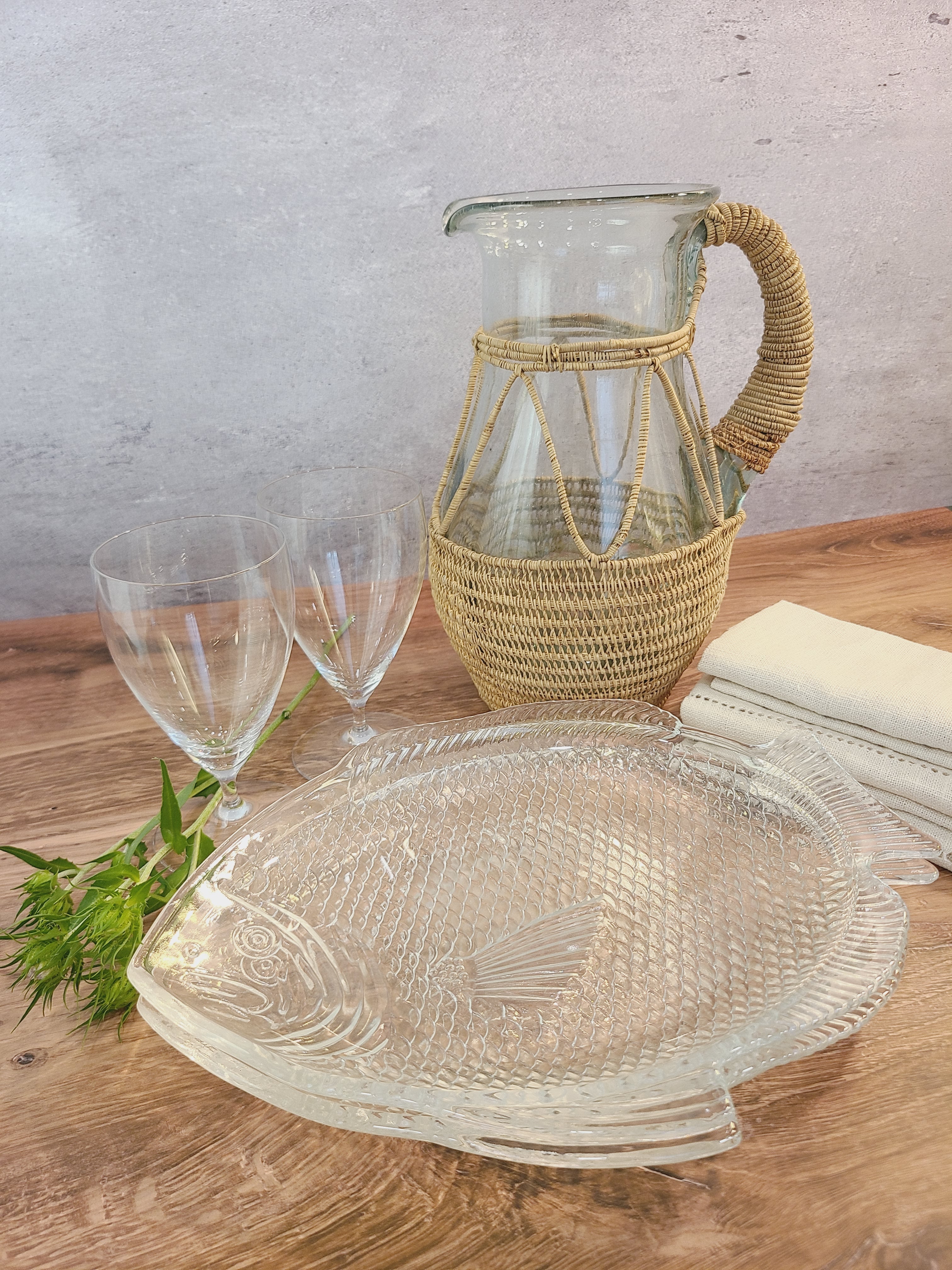 A vintage clear glass pitcher wrapped in a woven natural fiber displayed with clear glasses and clear glass fish plates.