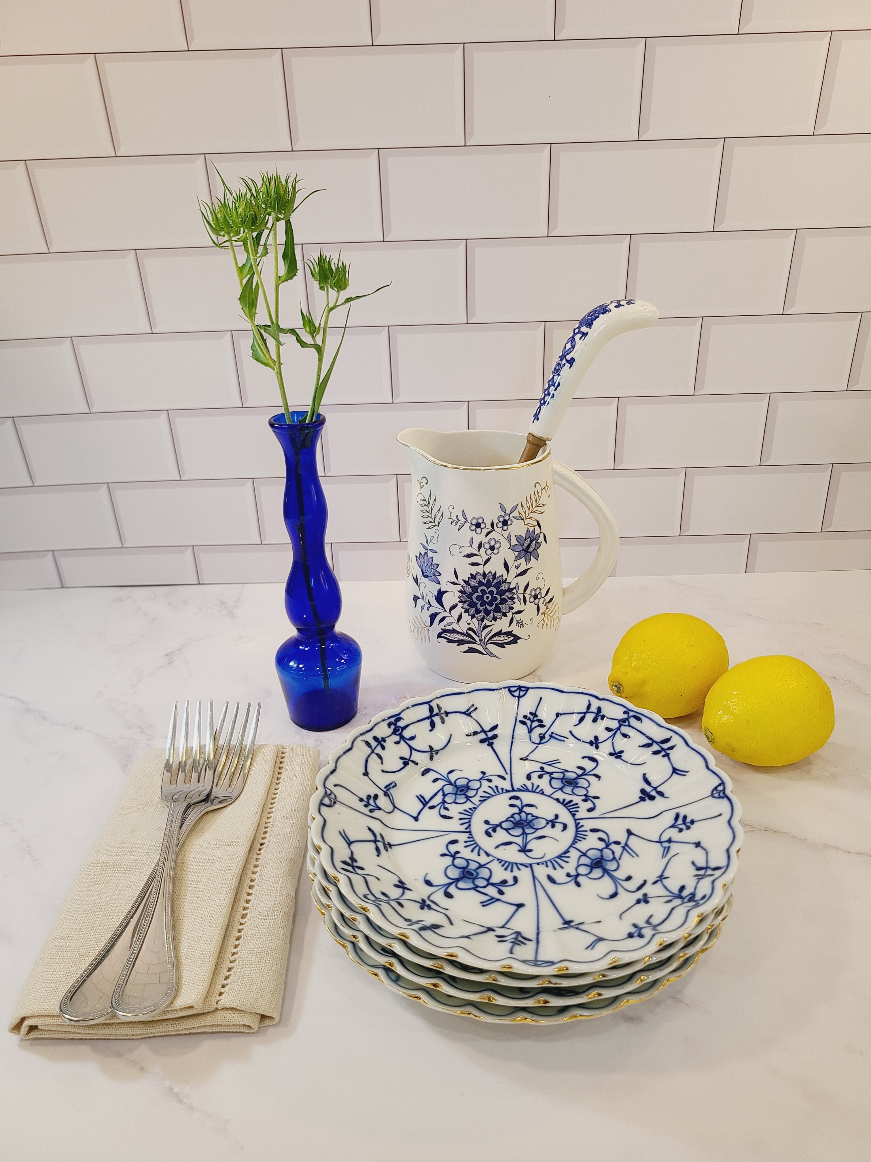 A vintage cobalt blue glass bud vase set in a styled tabletop arrangement featuring a stack of blue and white floral ceramic plates, a white pitcher with blue design and lemons against a white subway tile backsplash.