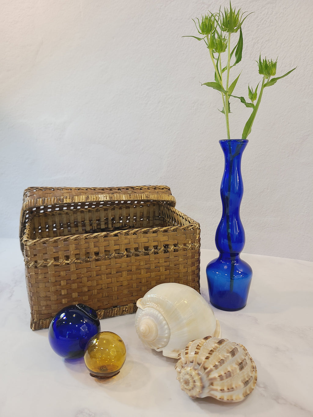 A vintage cobalt blue glass bud vase  styled with a woven basket, seashells, and fishing floats.