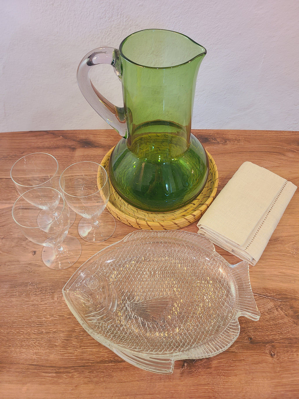 A vintage green glass pitcher with a clear handle, displayed on a woven coaster on a wooden table alongside a fish-shaped clear glass serving dish, four clear glasses, and a folded napkin.