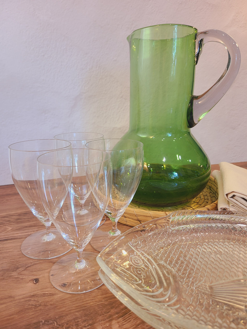 A vintage green glass pitcher with a clear handle sits on a wooden table alongside several clear wine glasses, a clear textured serving platter, and a folded cream napkin against a light wall.