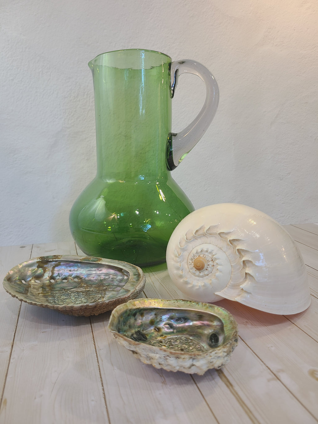 A vintage green glass pitcher styled with a large white seashell and two abalone shells on a light wood background.