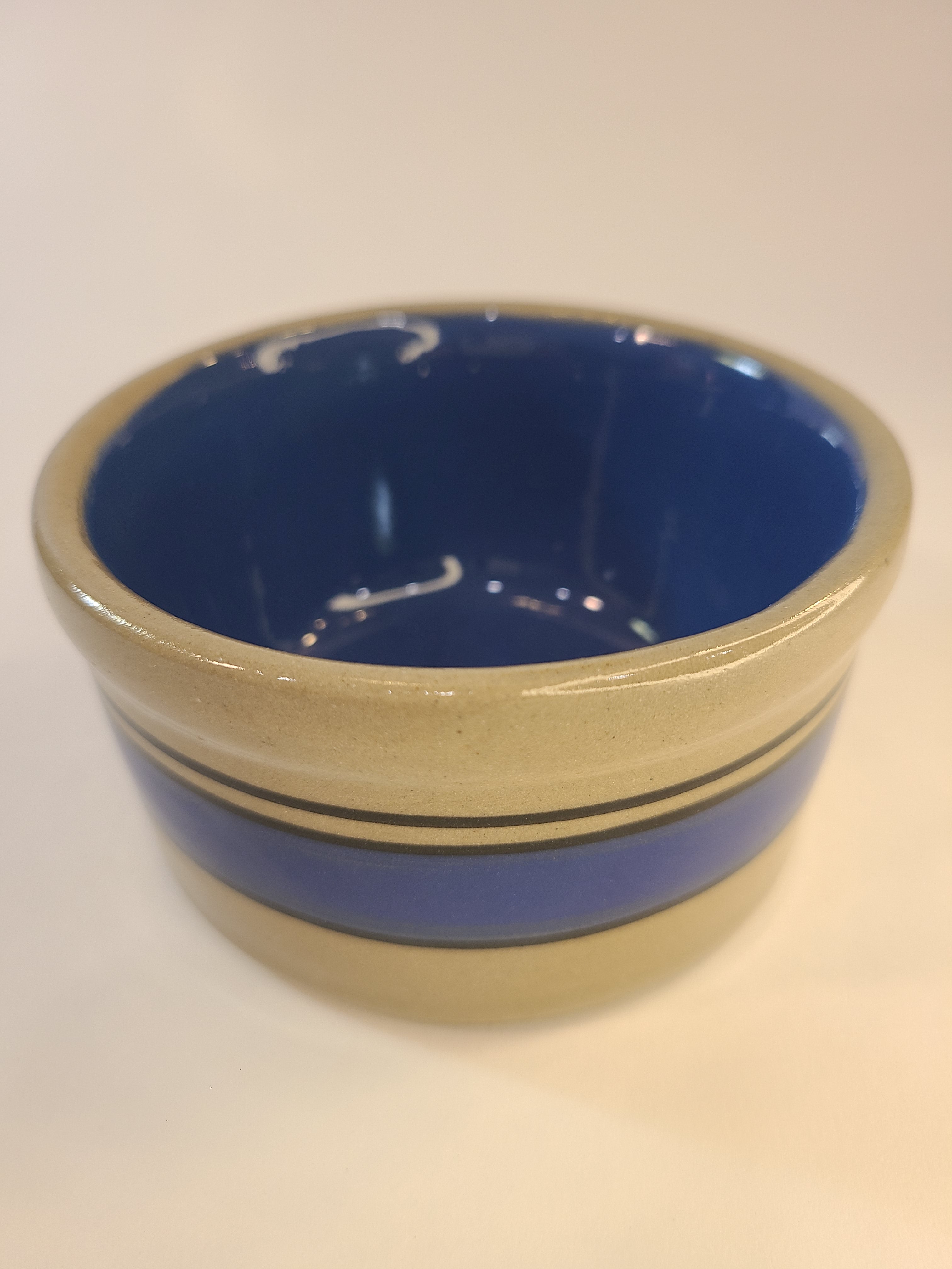 A small vintage stoneware crock with a deep blue interior and gray exterior accented by horizontal blue stripes.