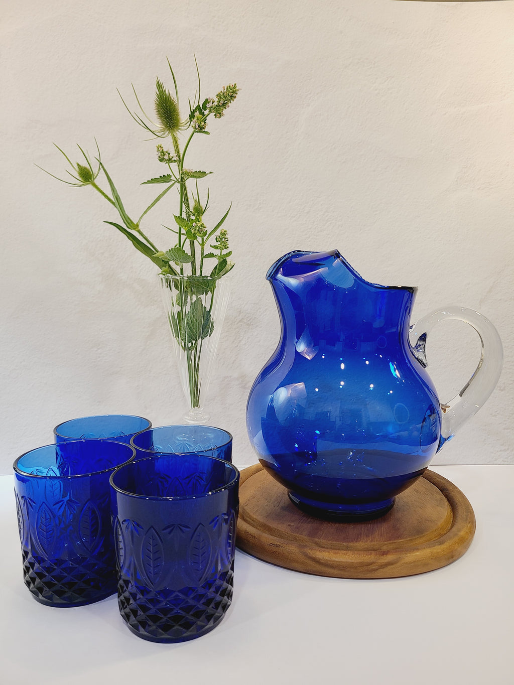 A cobalt blue glass pitcher with a clear glass handle, shown with cobalt blue glasses and a clear glass vase with greenery.