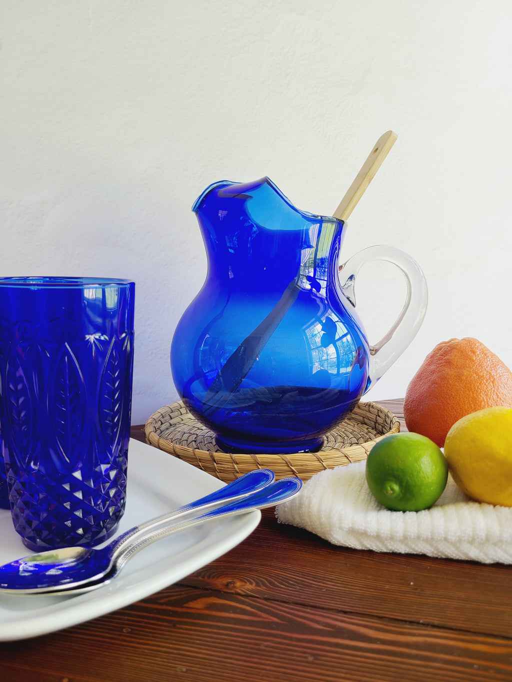 A vintage cobalt blue glass pitcher with a clear glass handle holding a wooden spoon shown with citrus fruit, cobalt blue glasses, and silverware.