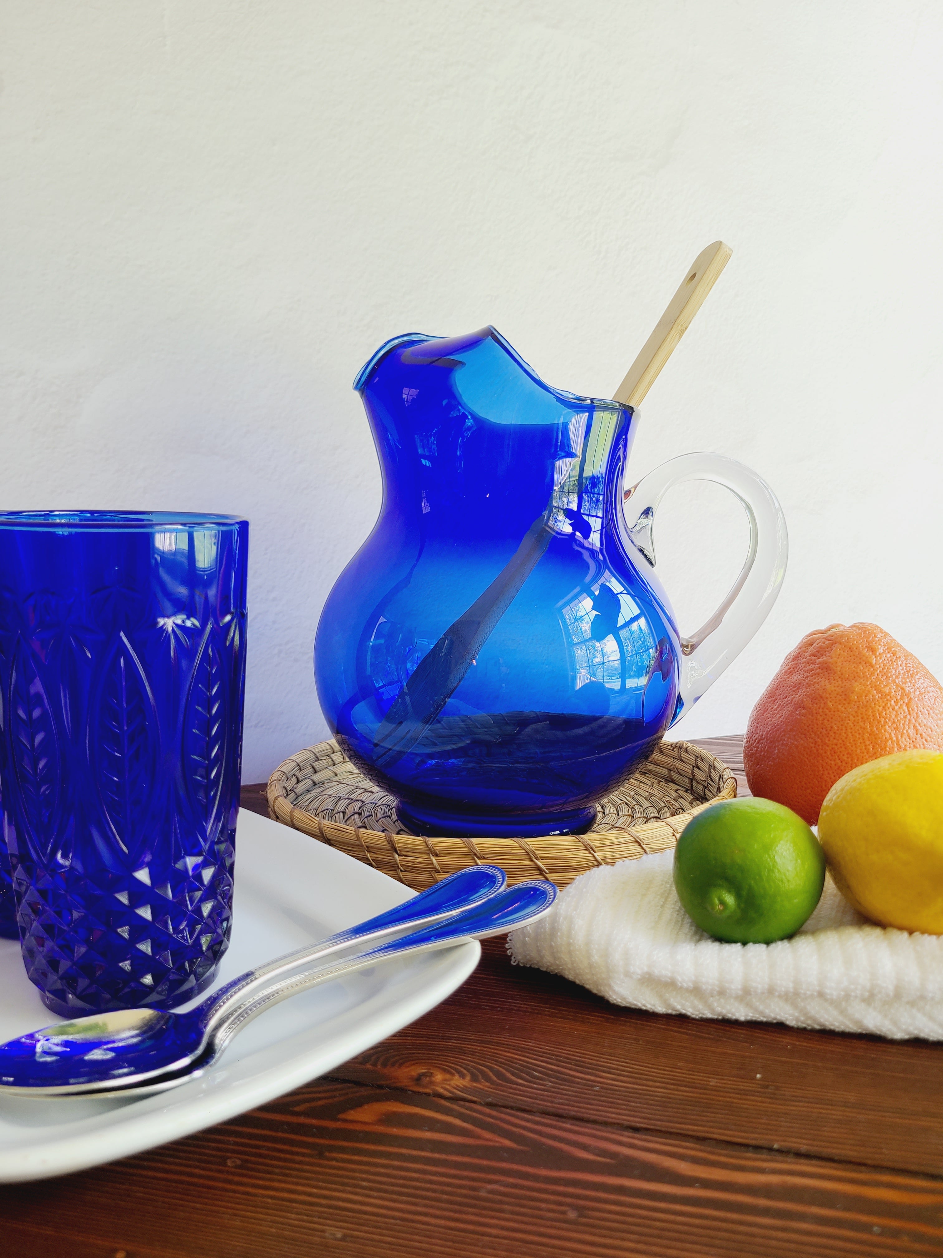 A vintage cobalt blue glass pitcher with a clear glass handle holding a wooden spoon shown with citrus fruit, cobalt blue glasses, and silverware.