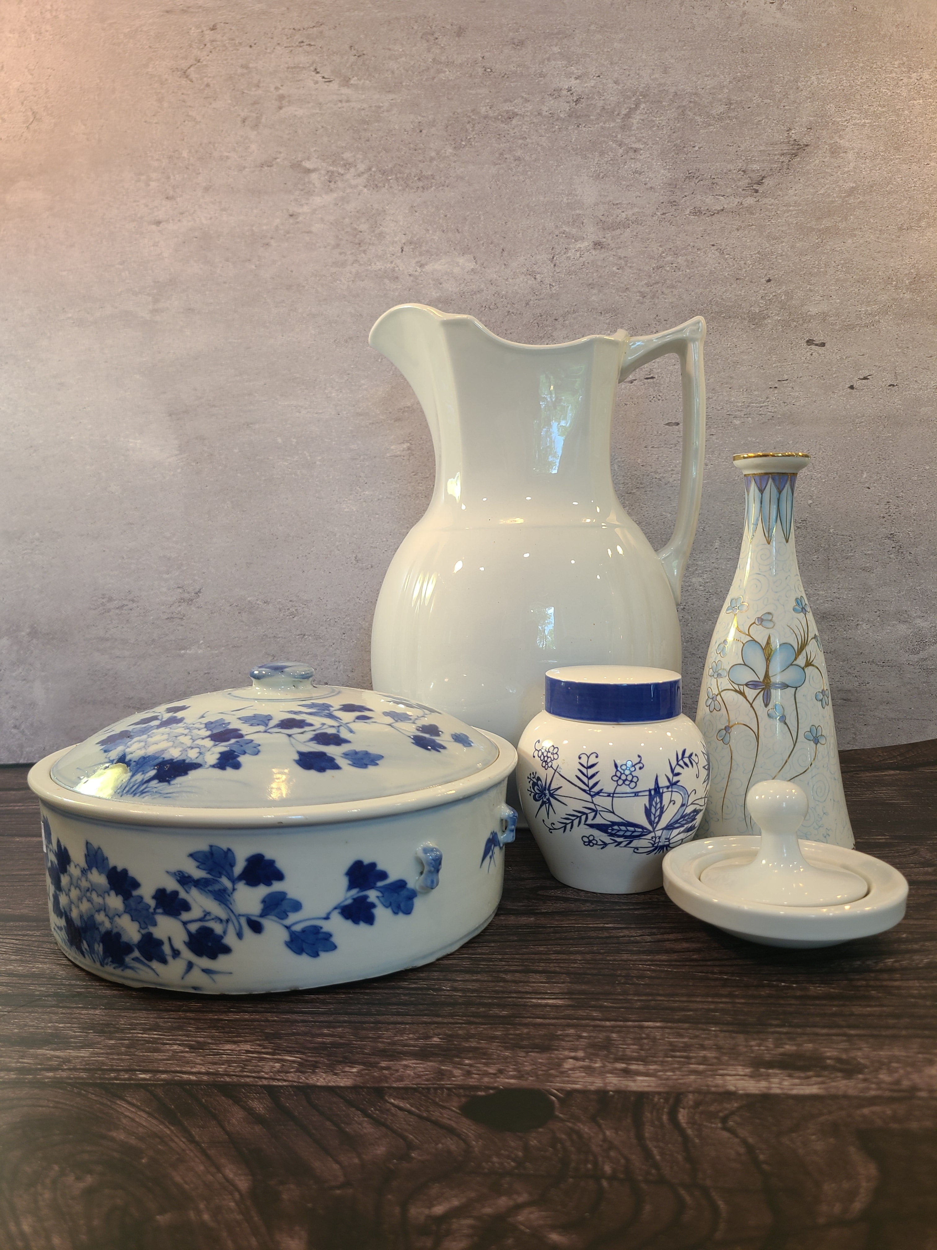 A vintage Japanese white ceramic tea jar with a blue floral hand-painted pattern and a blue and white lid displayed on a dark wood table with other ceramic pieces.