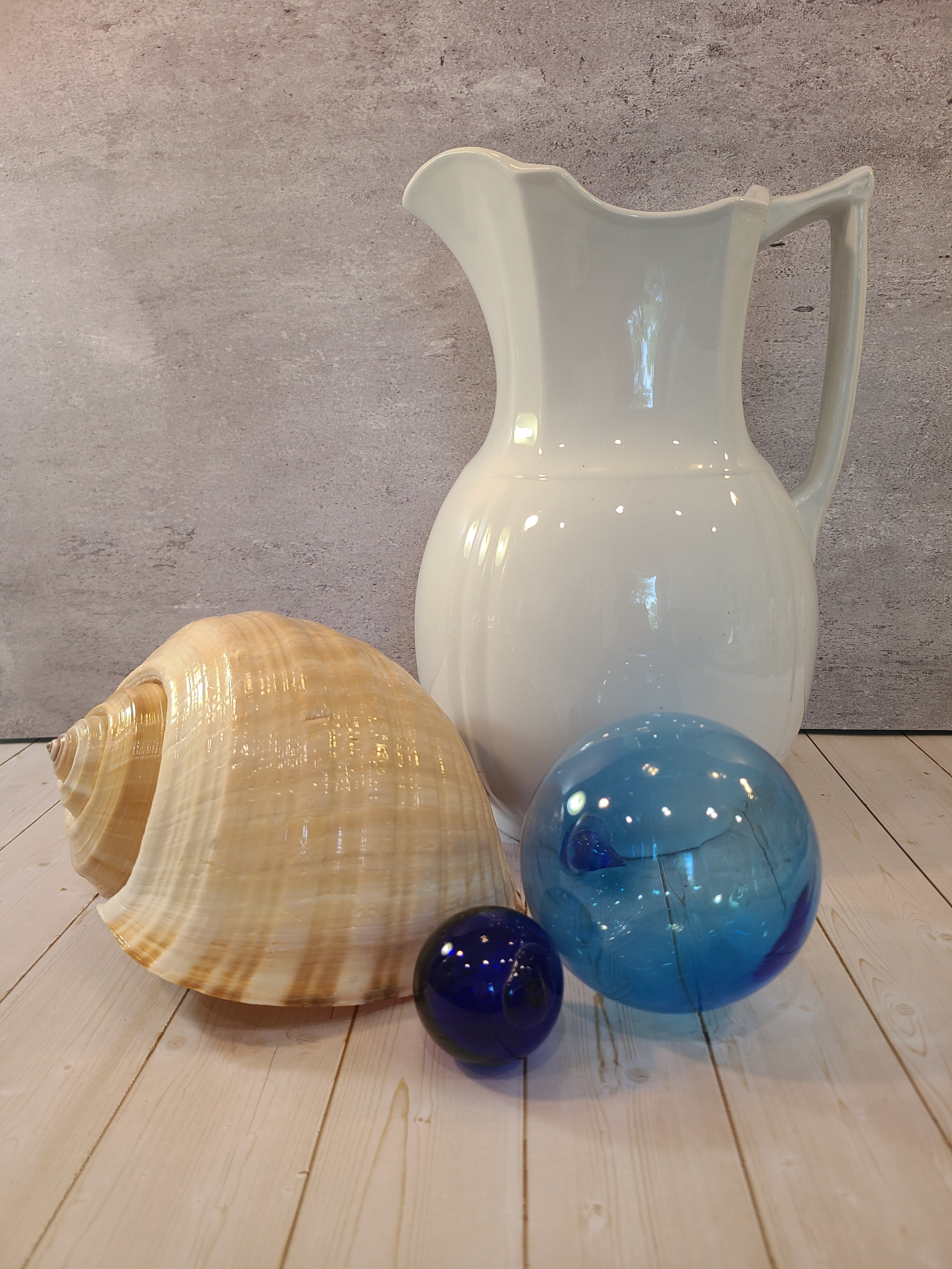A large tun seashell displayed with glass fishing floats and a white ironstone pitcher.