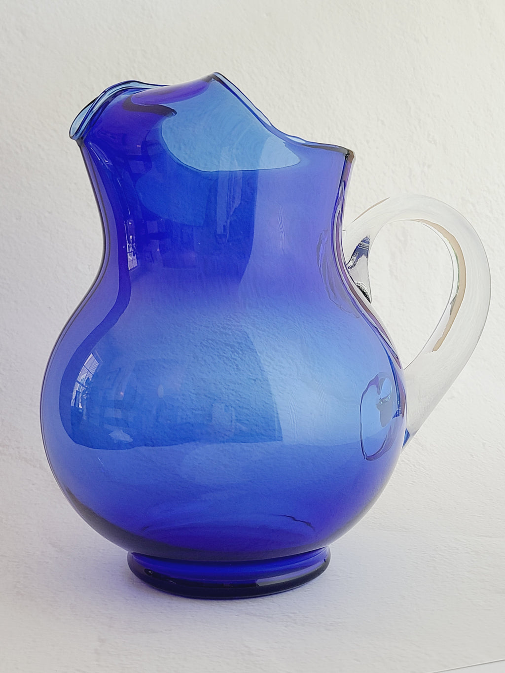 A vintage cobalt blue glass pitcher with a clear glass handle, shown against a plain white wall.