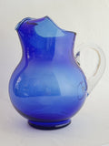 A vintage cobalt blue glass pitcher with a clear glass handle, shown against a plain white wall.
