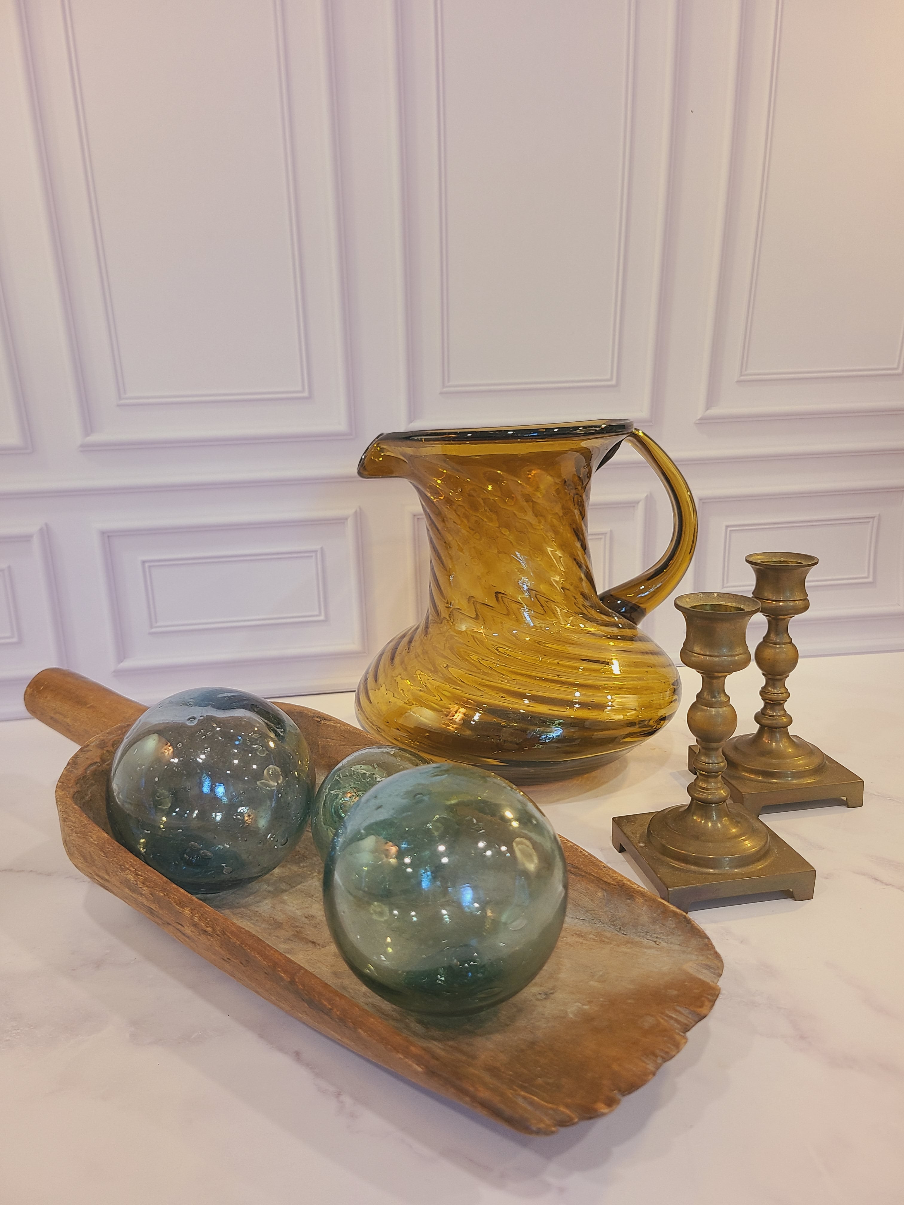 A pair of vintage brass candle holders displayed with amber glass pitcher and wooden scoop holding authentic glass fishing floats.