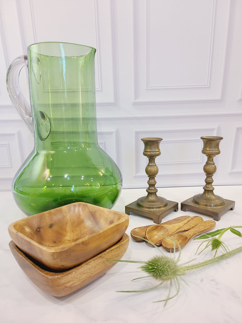 A pair of vintage brass candle holders displayed with green glass pitcher, wooden bowls and spoons on white table.
