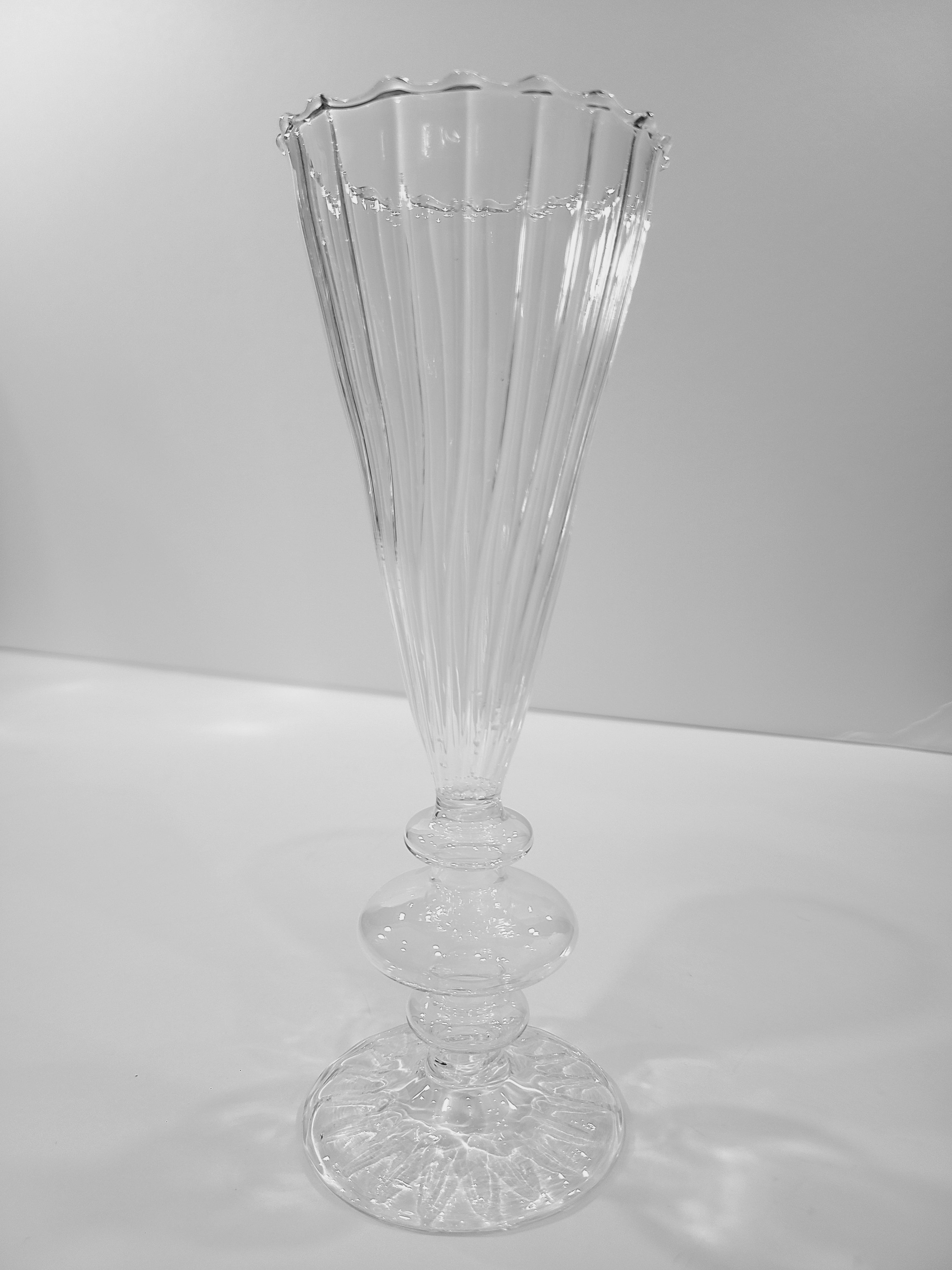 A clear ribbed glass vase with a flared, scalloped rim and a pedestal base, photographed against a plain light gray background.