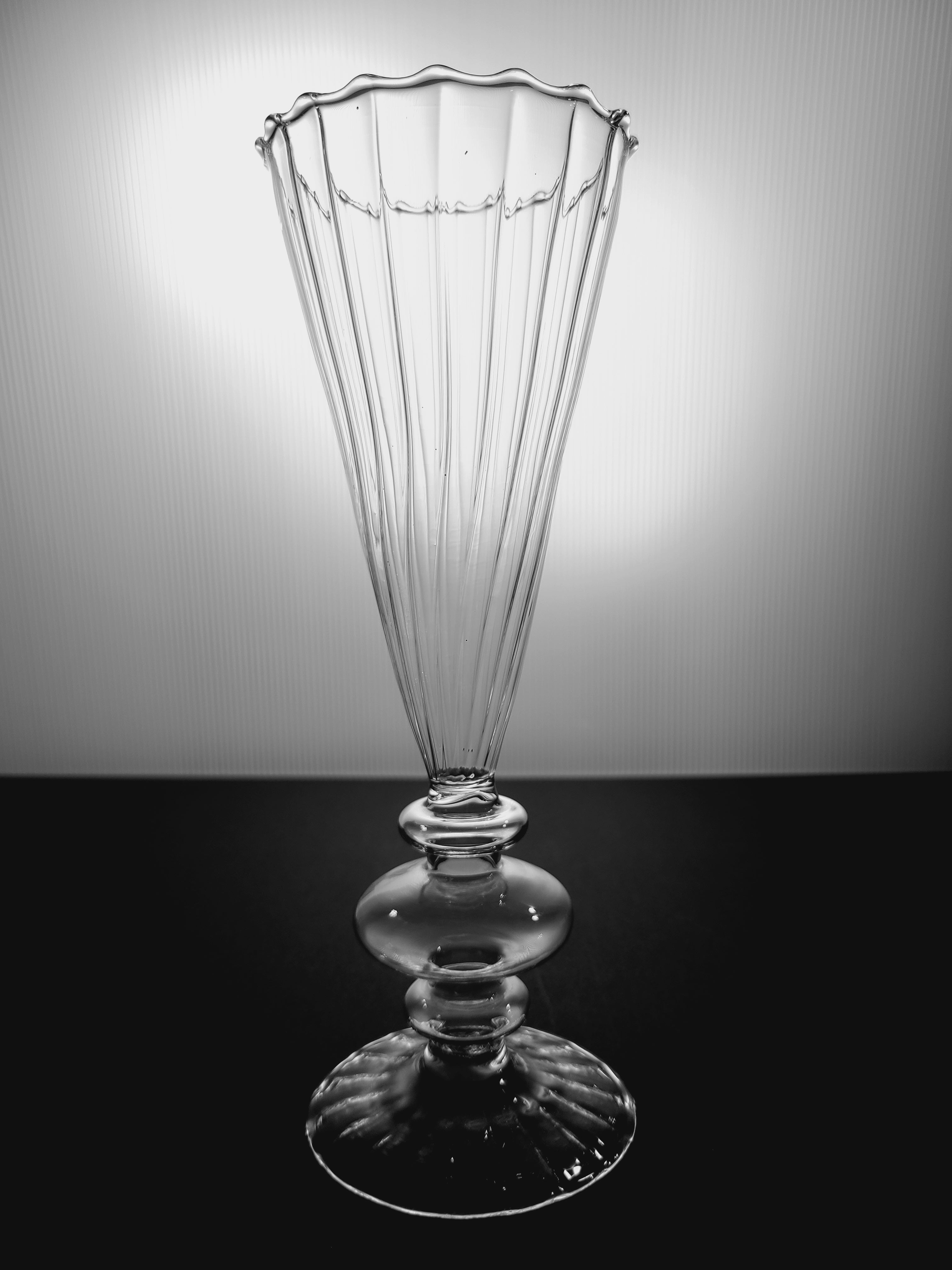 A clear ribbed glass vase with a flared, scalloped rim and a pedestal base, backlit and photographed on a dark base.