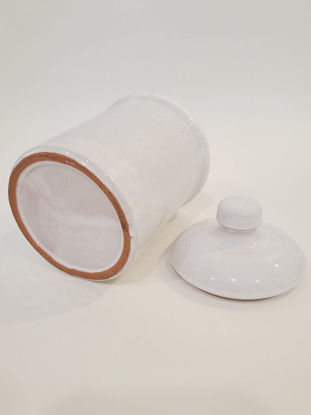 A white ceramic crock with a matching lid, shown on a plain white background. The jar is tipped on its side to reveal a light brown clay rim bottom, and the lid sits next to it.