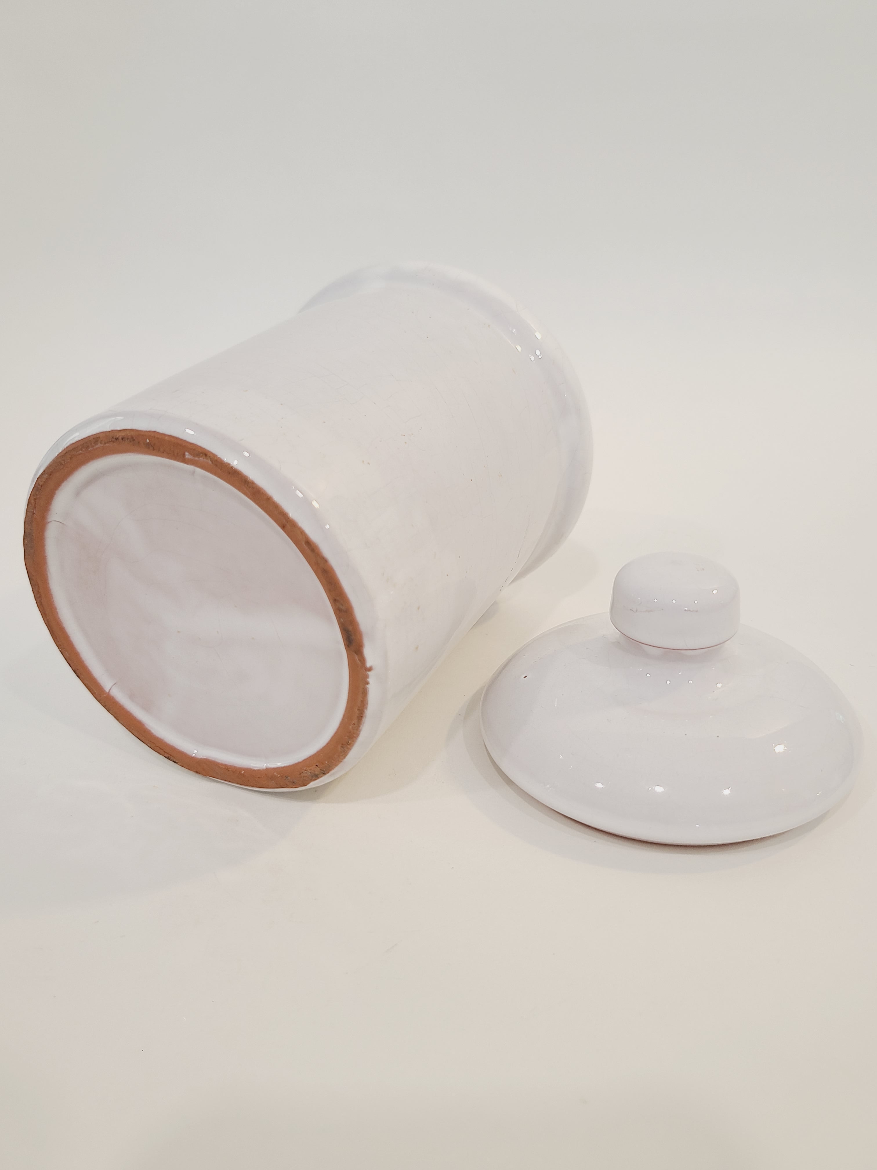 A white ceramic crock with a matching lid, shown on a plain white background. The jar is tipped on its side to reveal a light brown clay rim bottom, and the lid sits next to it.