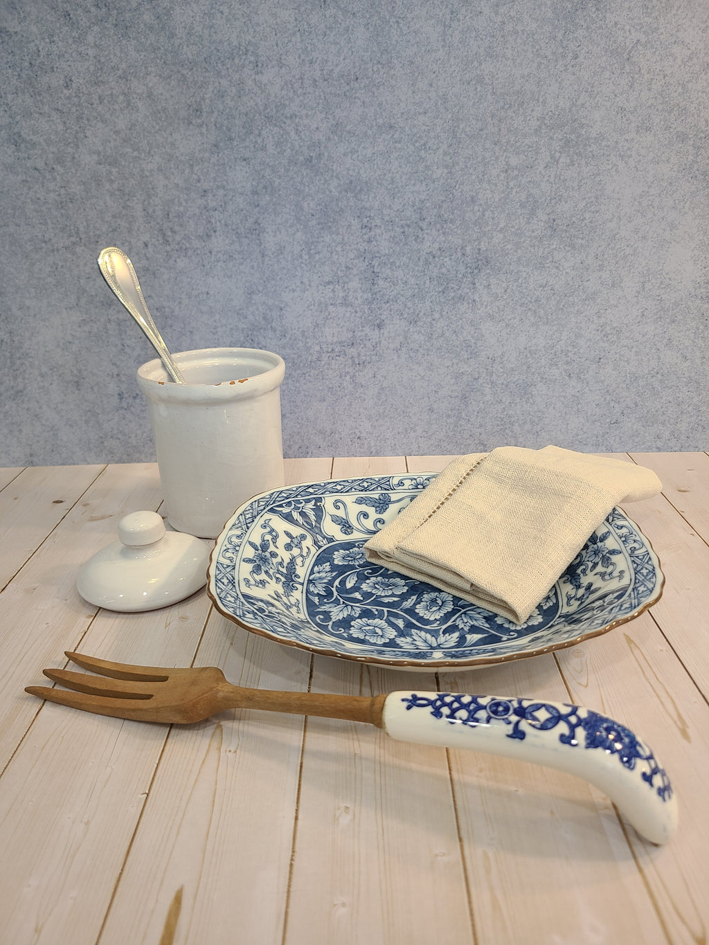 A styled tabletop arrangement featuring a white ceramic crock with a  spoon, a white ceramic plate with blue floral patterns and a folded cream napkin, and wood and ceramic fork, set against a split gray and light wood plank background.