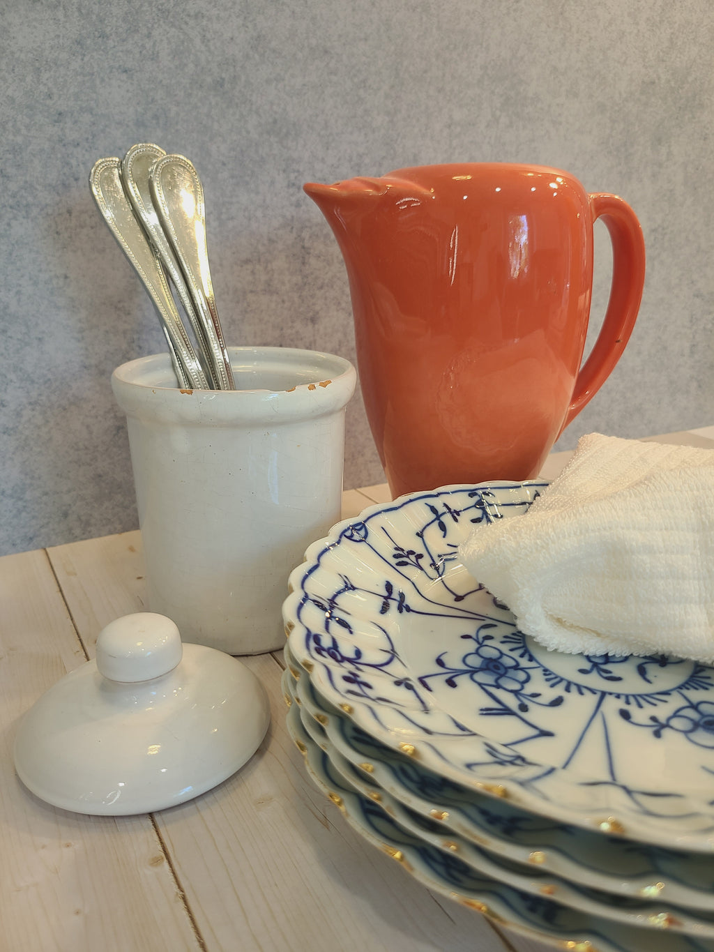 A collection of ceramic tableware including a glossy coral colored pitcher, a white crock with spoons, a stack of blue and white patterned plates with gold trim, and a white cloth napkin on a light wood surface.