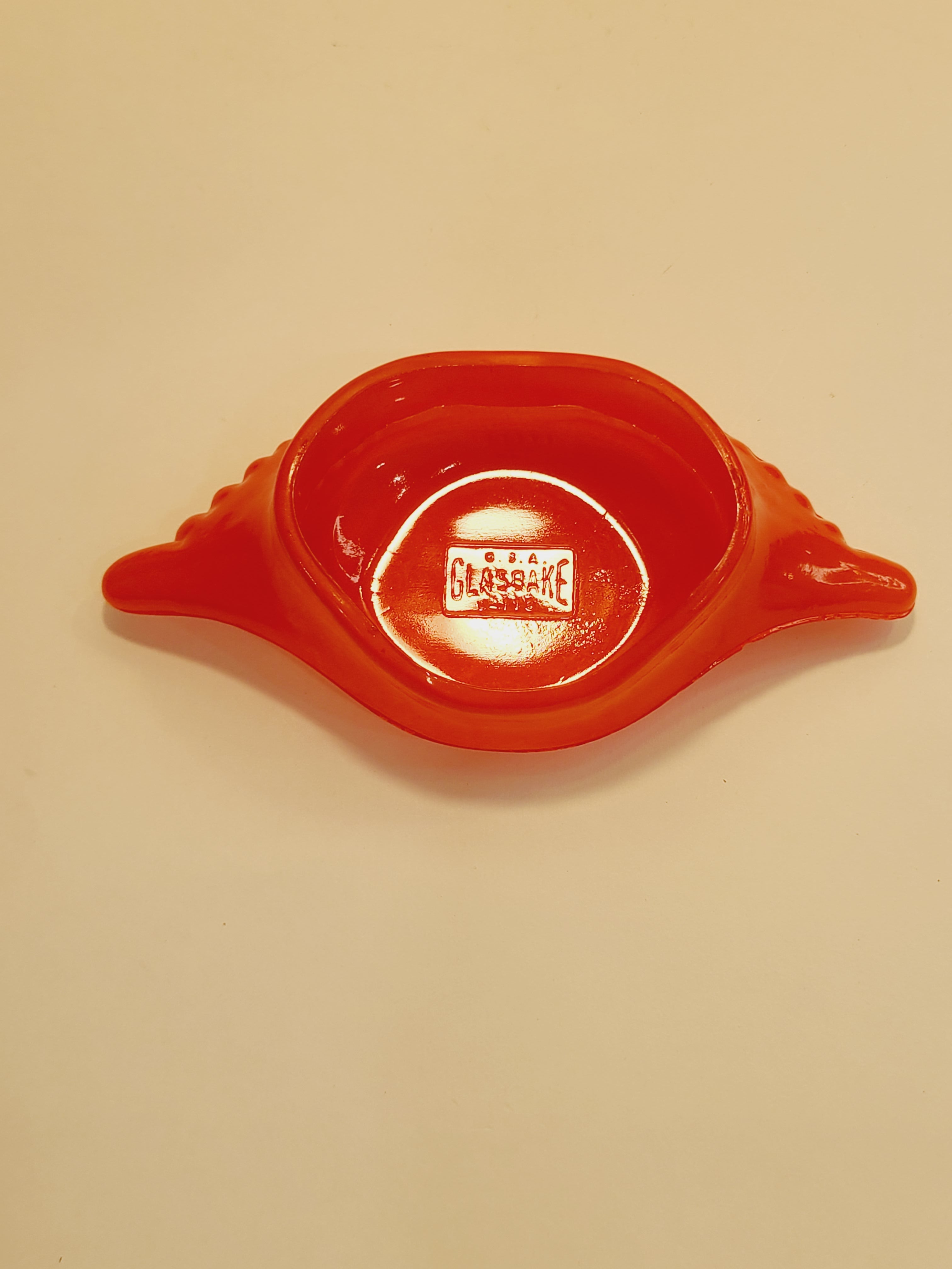 Red-orange Glasbake glass dish shaped like a crab shell, 5.75 inches by 2.75 inches showing logo pressed into glass.