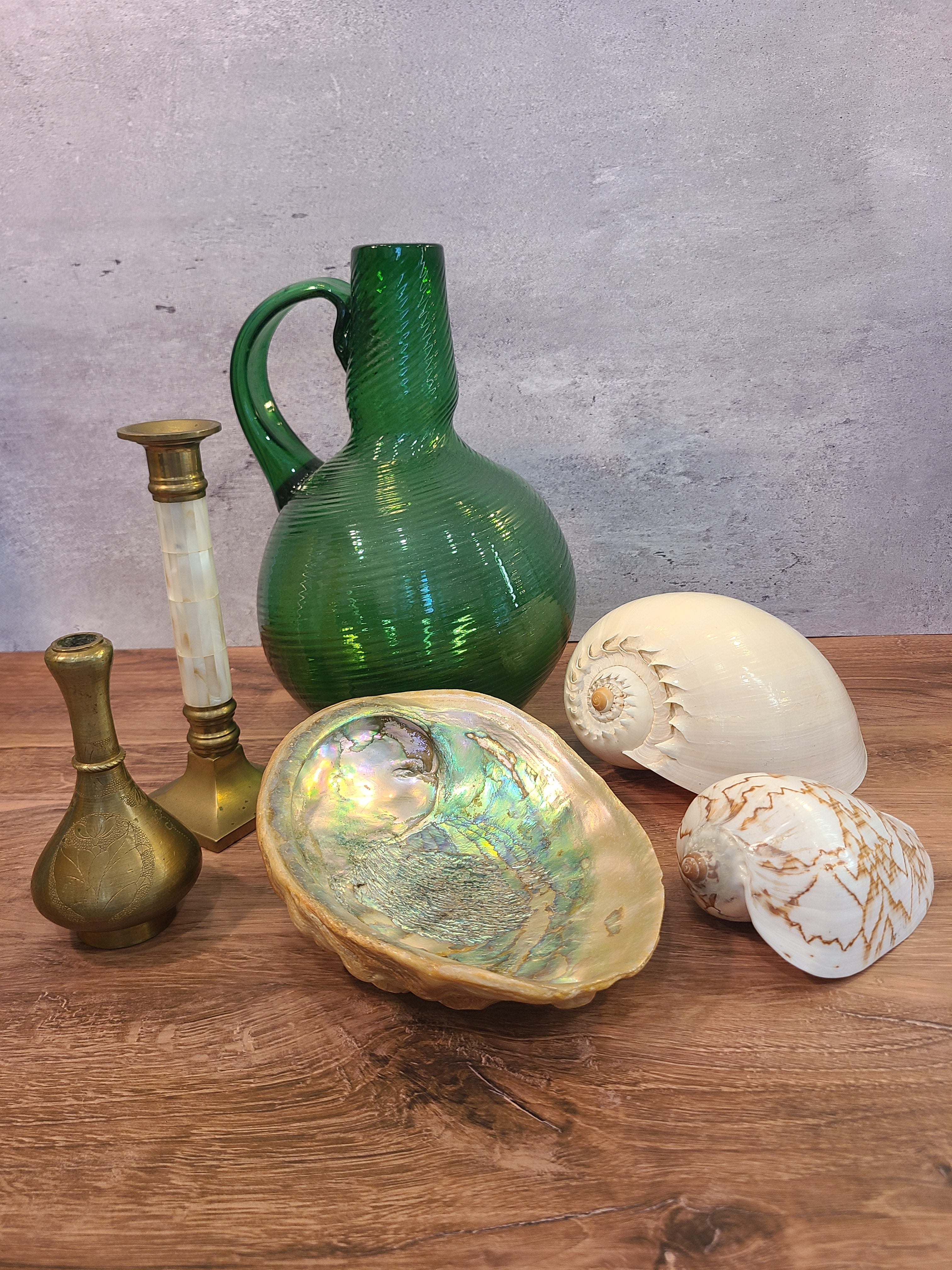 Natural abalone shell specimen, iridescent rainbow colors, 8.25 inches long by 6.5 inches wide, displayed with green glass jug, other shells, brass vase and candle holder on wood surface.