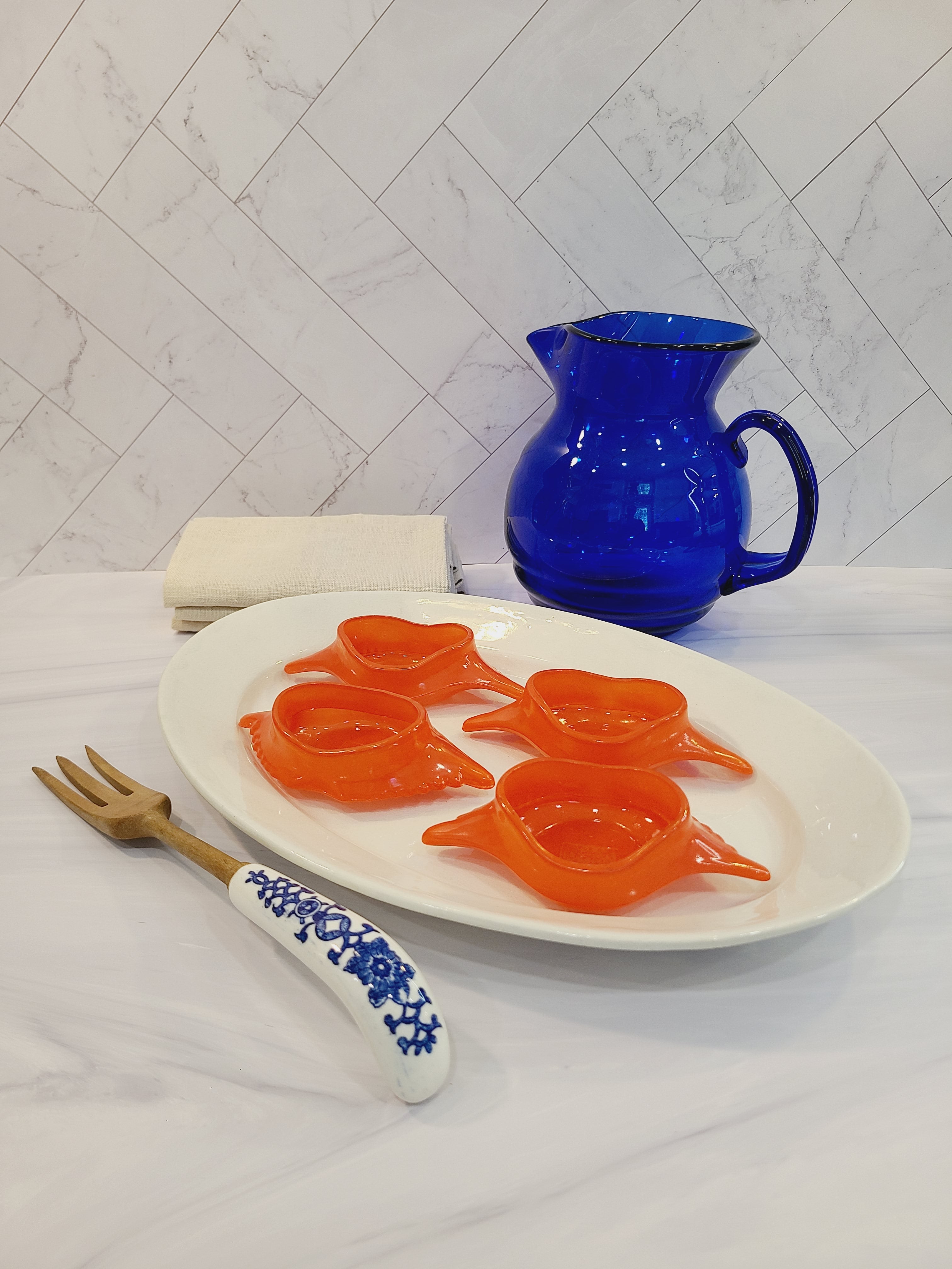 Red-orange Glasbake glass dishes shaped like a crab shell, 5.75 inches by 2.75 inches displayed on a white platter with a cobalt blue pitcher and wooden utensil with porcelain handle.