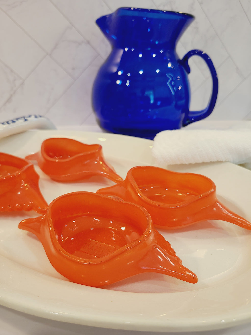 Close up of Red-orange Glasbake glass dishes shaped like a crab shell, 5.75 inches by 2.75 inches displayed on a white platter with a cobalt blue pitcher in the background.