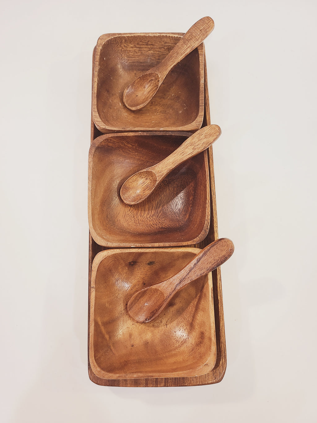 Overhead view of mid-century monkey pod wood condiment serving set, three square bowls with carved wooden spoons on rectangular tray
