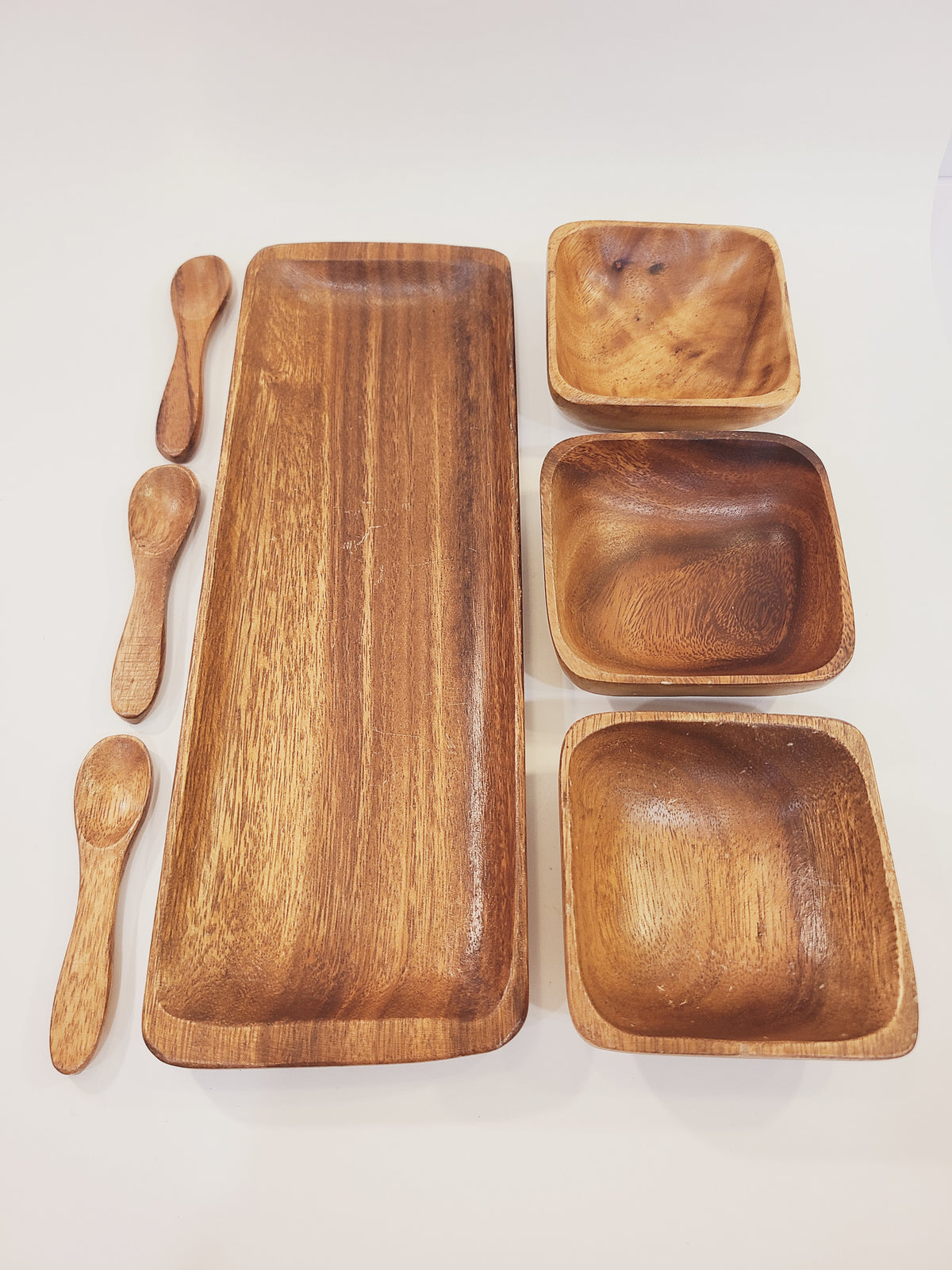 Flat lay of complete 7-piece monkey pod wood serving set showing rectangular tray, three square bowls, and three carved spoons, handcrafted Philippines 1950s–1960s