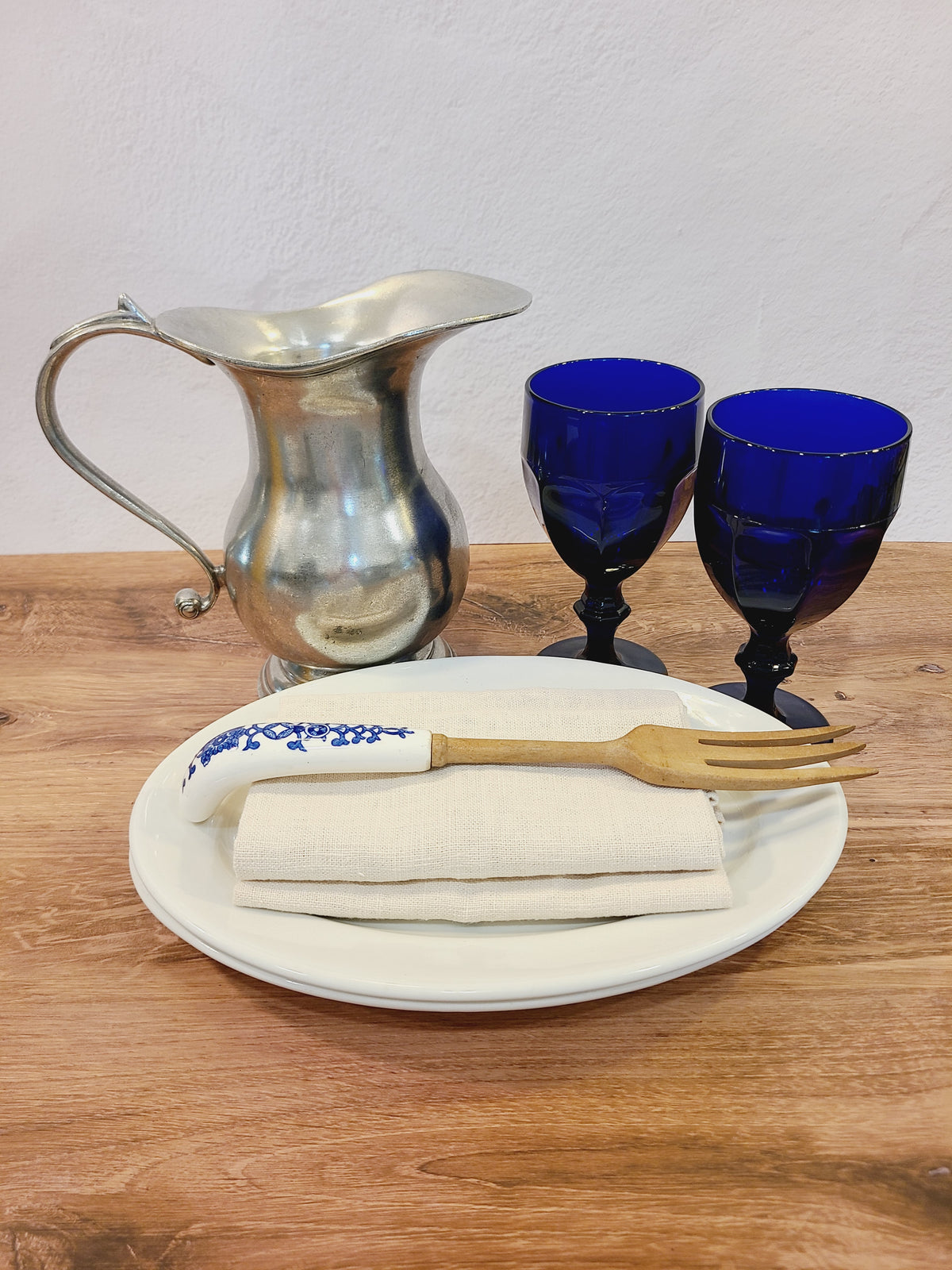 Libbey Gibraltar cobalt blue goblets styled with pewter pitcher, linen napkin, and brass fork on wood table, elegant coastal vintage dining setting