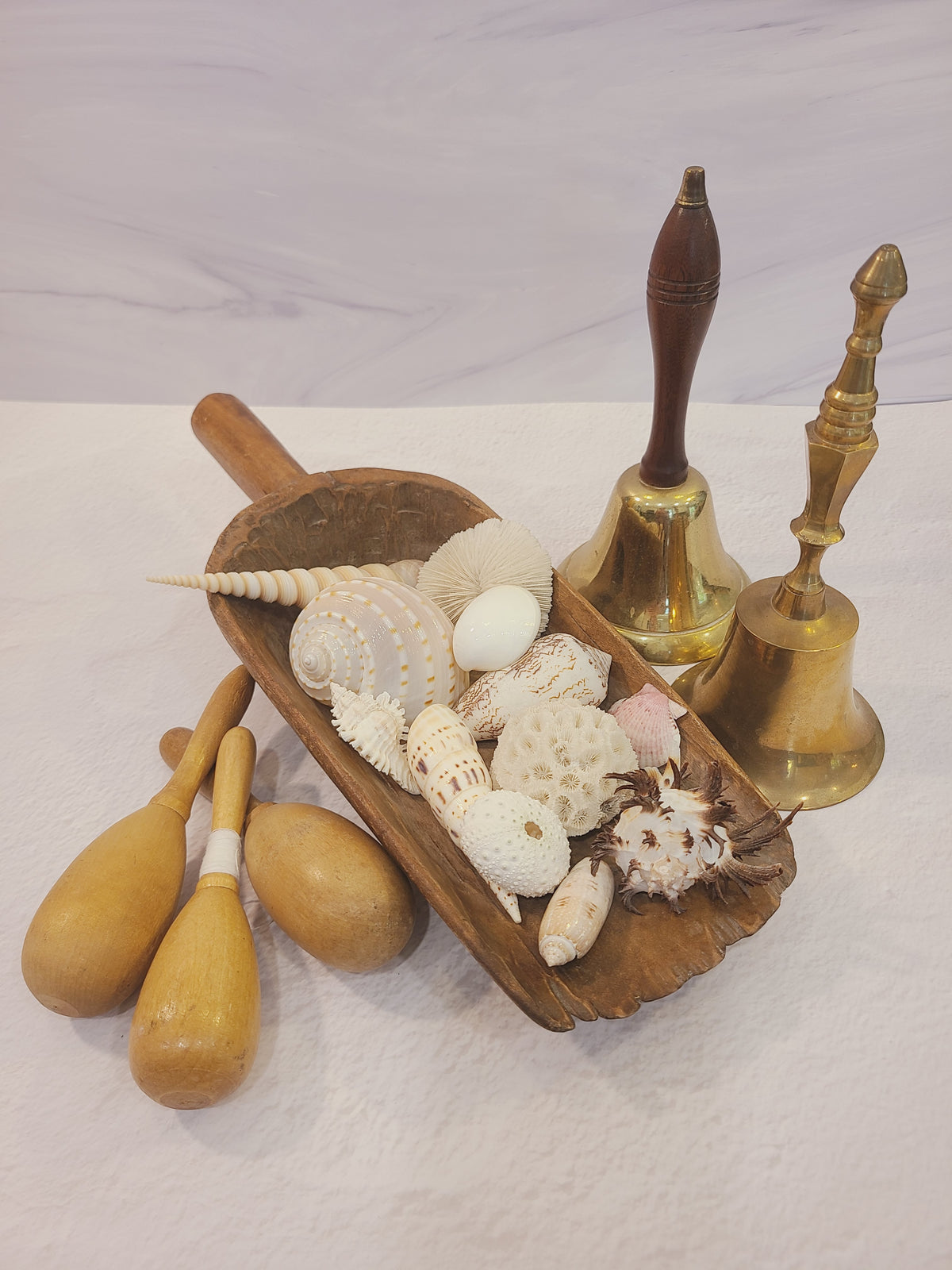 Antique hardwood grain scoop styled with shells, brass hand bells, and wooden pins — coastal farmhouse vignette decor