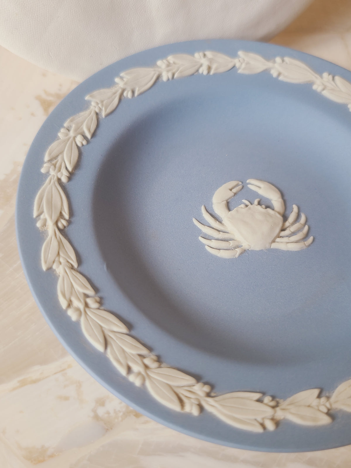 Angled close-up of Wedgwood blue jasperware crab pin plate showing white bas-relief Cancer zodiac cameo and laurel wreath border, coastal blue and white decor