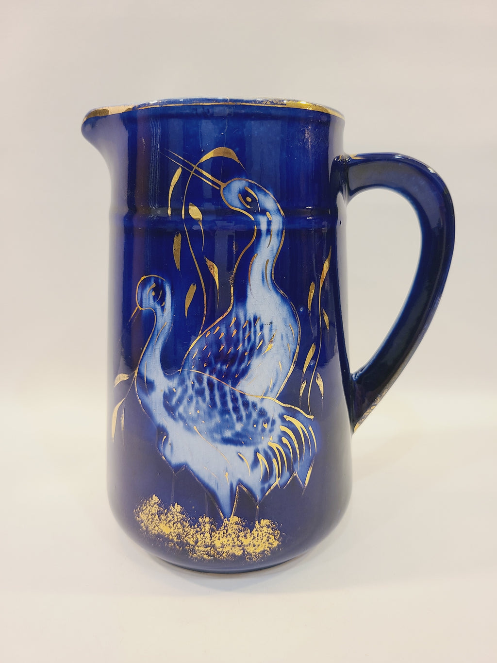 Cobalt blue ceramic pitcher with hand-painted cranes, gold foliage, and gold-trimmed rim