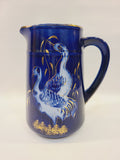 Cobalt blue ceramic pitcher with hand-painted cranes, gold foliage, and gold-trimmed rim